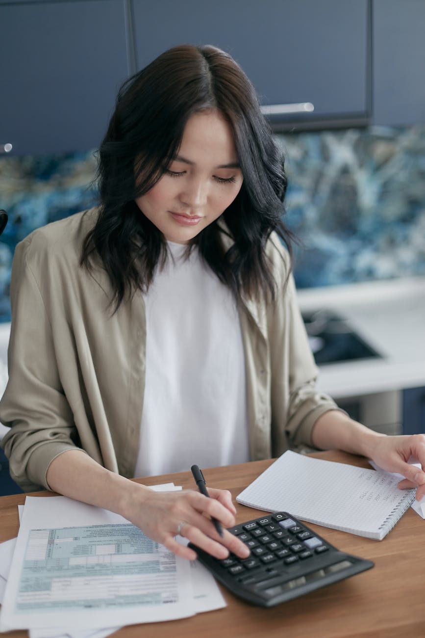 A woman at a wooden desk reviewing her household budget with a calculator and notebook in soft daylight